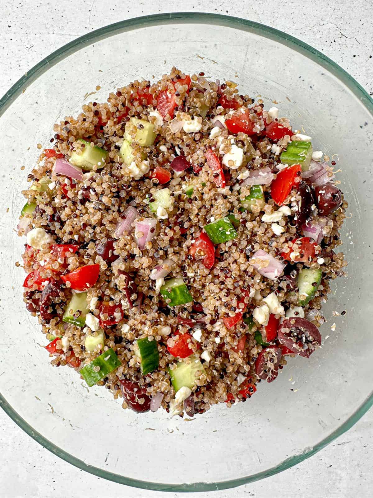 A clear bowl filled with tricolor quinoa salad tossed with cucumbers, red onions, cherry tomatoes, olives, feta, and vinaigrette.