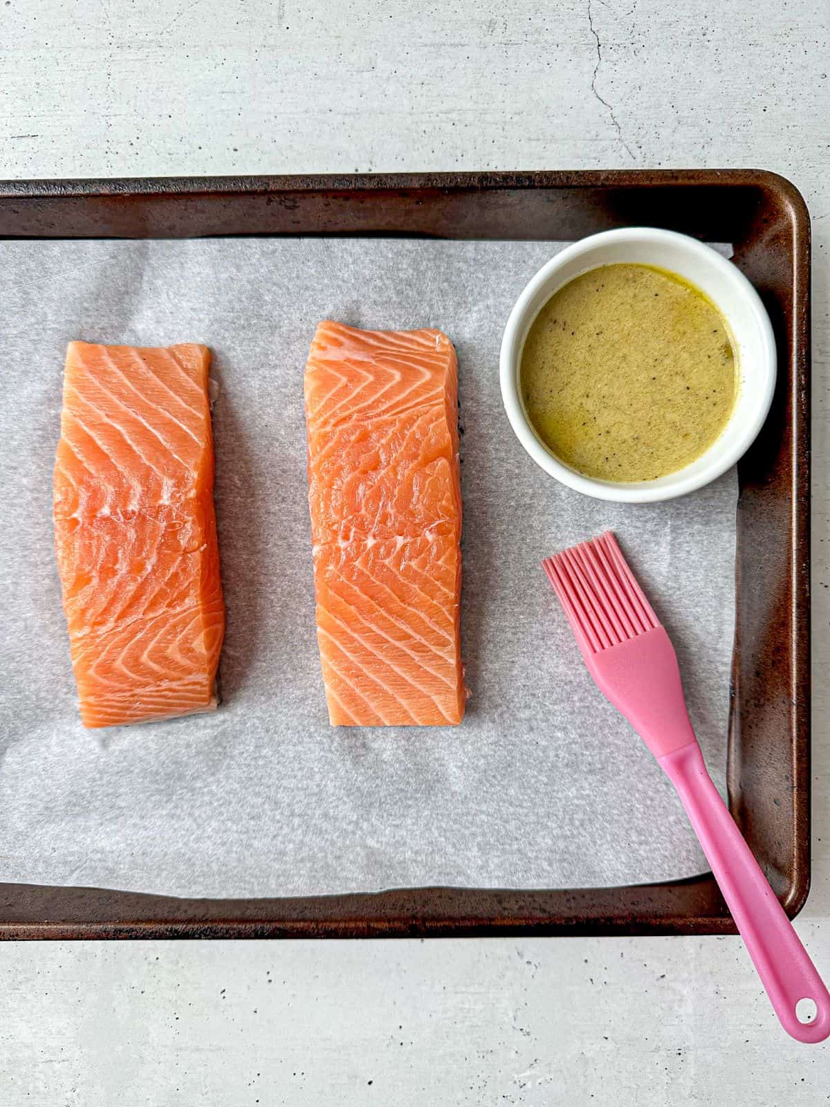 Two salmon fillets on a baking sheet covered with parchment paper. A small bowl of marinade and a pastry brush are off to the side.