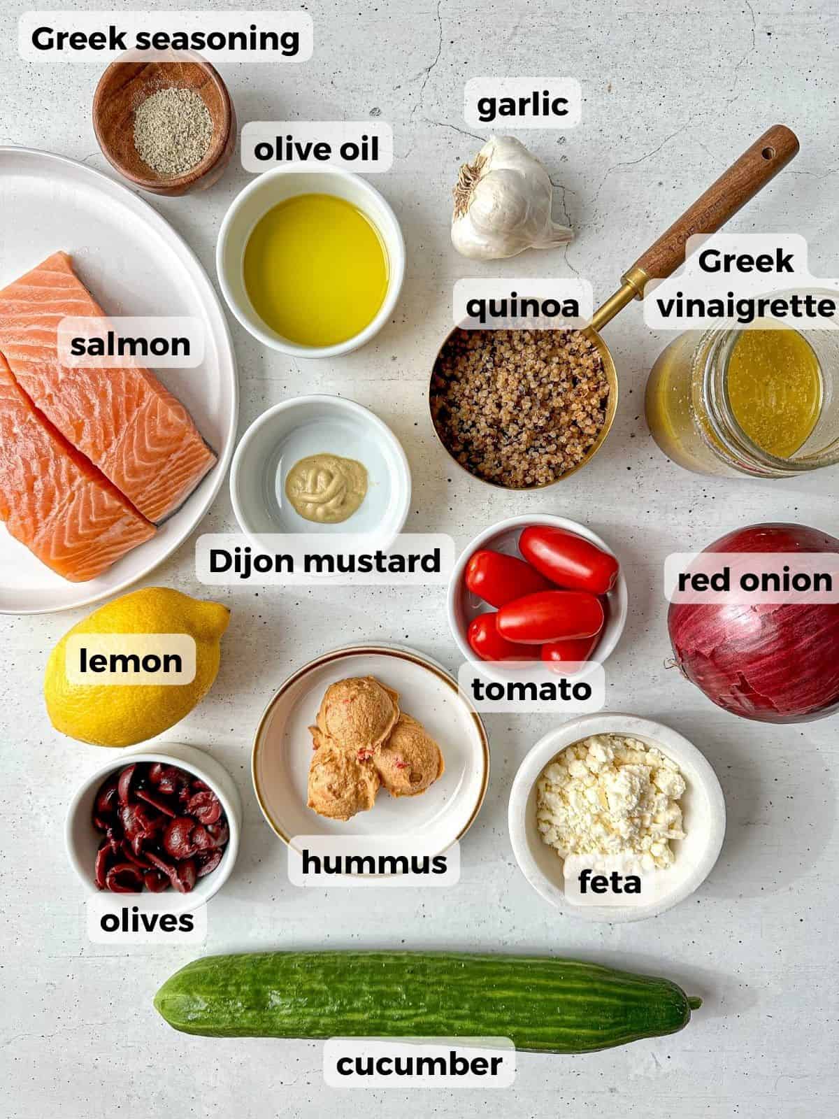 Ingredients needed to make this recipe are on a concrete counter. Ingredients include: salmon and quinoa, fresh veggies, feta cheese, olives, lemon, Dijon mustard, and Greek vinaigrette.