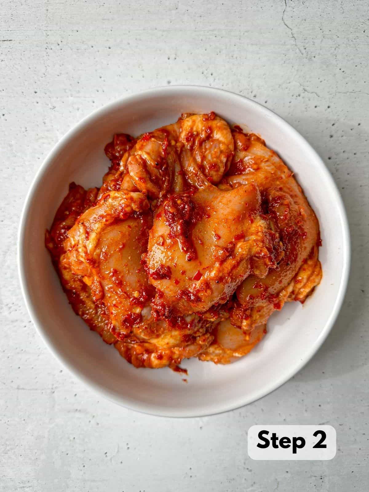 Chicken thighs coated in a red-orange harissa marinade in a white bowl.