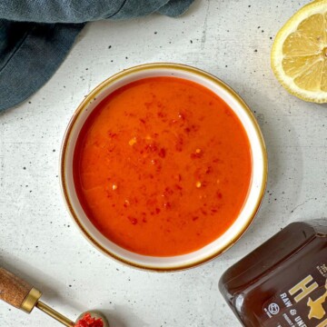 Orange-red colored hot harissa vinaigrette in a white bowl surrounded by a sliced lemon, jar of honey, and measuring spoon of harissa paste.