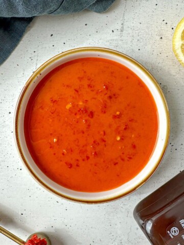 Orange-red colored hot harissa vinaigrette in a white bowl surrounded by a sliced lemon, jar of honey, and measuring spoon of harissa paste.