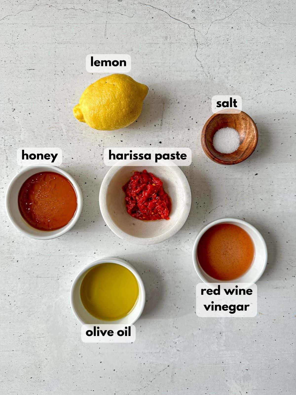 Ingredients needed to make this recipe labeled and spread out on a countertop: harissa paste, olive oil, lemon, red wine vinegar, honey, and salt.