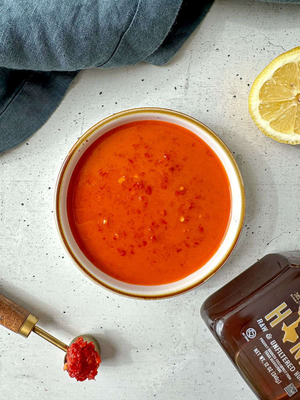 Orange-red colored harissa vinaigrette in a white bowl surrounded by a sliced lemon, jar of honey, and measuring spoon of harissa paste.