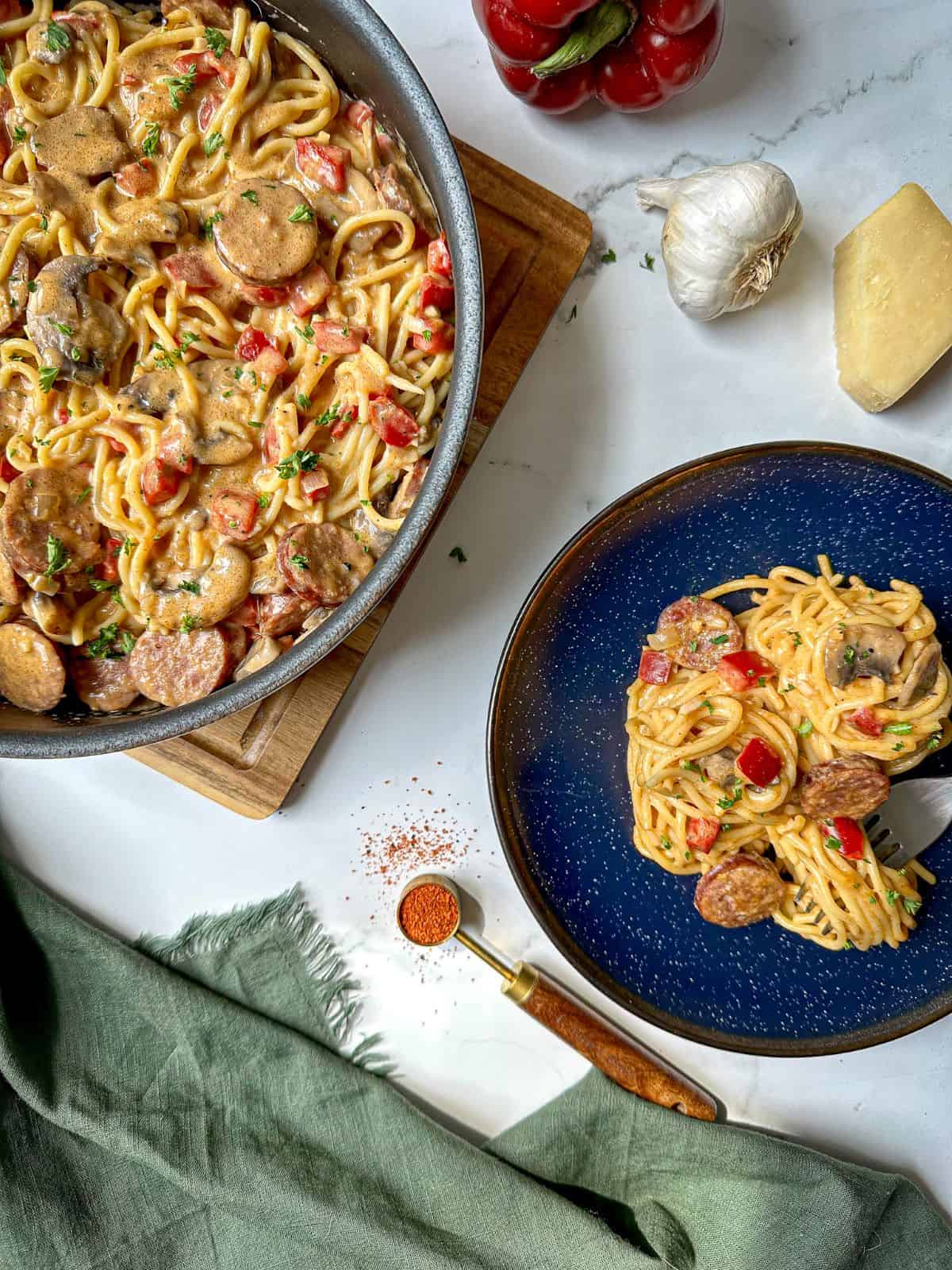 A skillet of Cajun spaghetti with sausage, peppers, and mushrooms next to a blue plate with a serving size and fork.