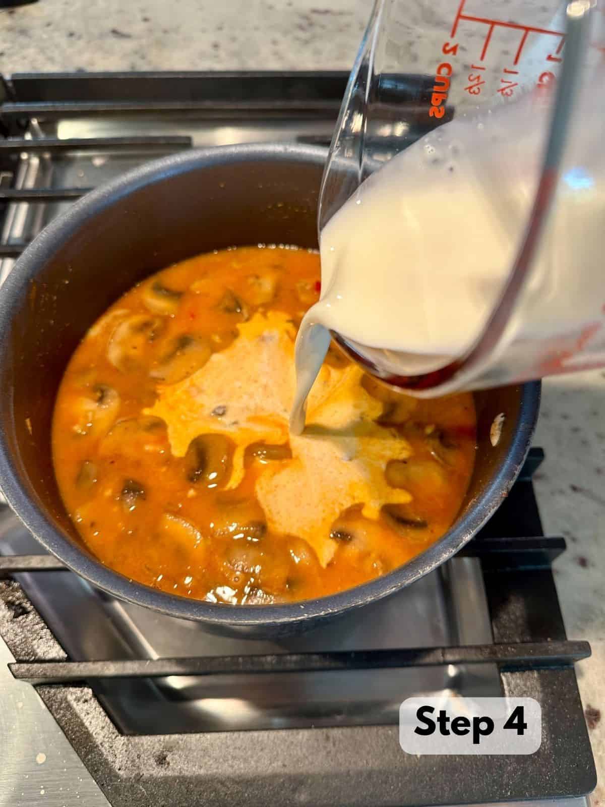 A measuring cup of heavy cream being poured into a saucepot on a stovetop.