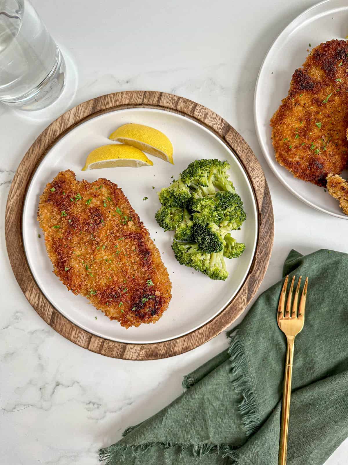 A plate with panko fried chicken, broccoli, and lemon with a fork and green towel on the side.