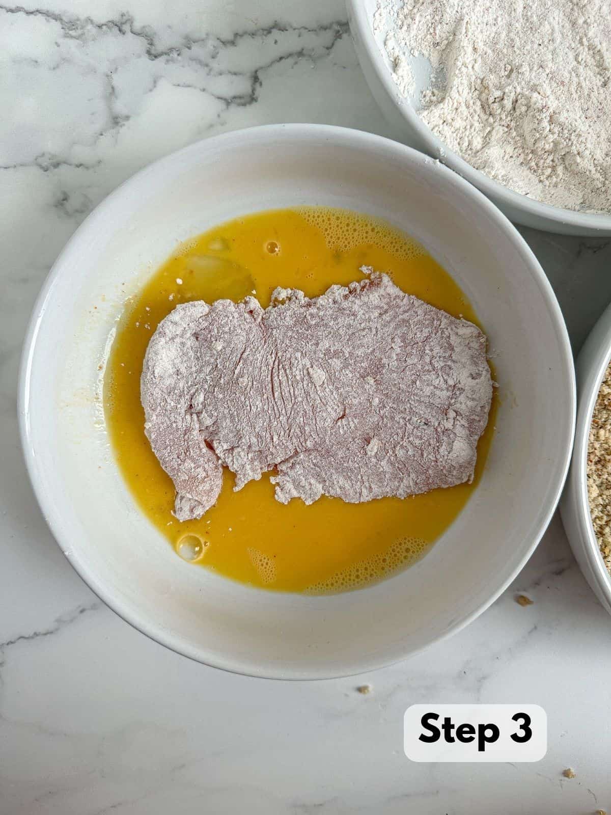 A bowl with a whisked egg conatins an uncooked chicken breast that is coated in flour.