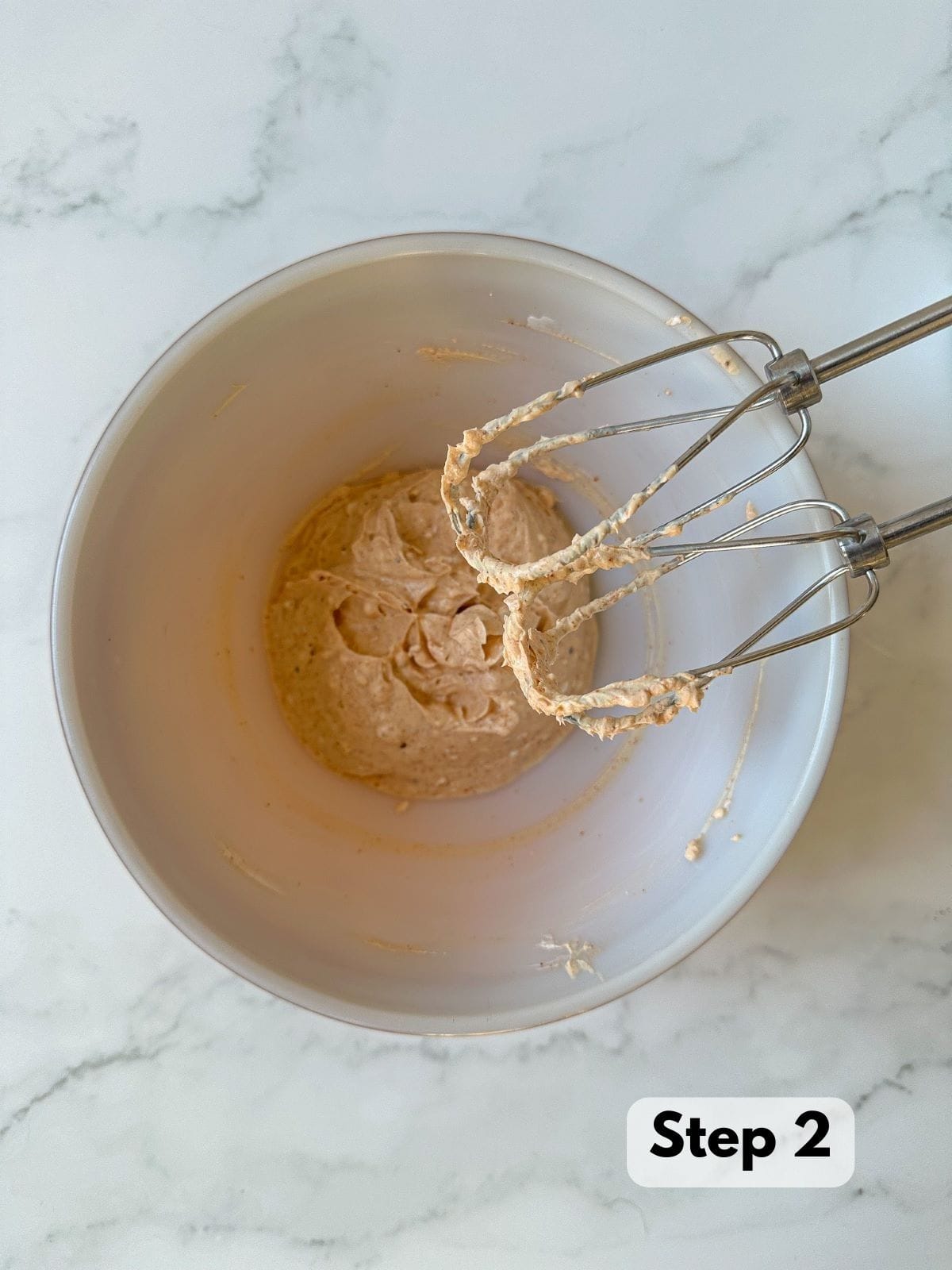 A hand mixer next to a mixing bowl of blended cream cheese, sour cream, hot sauce, seasonings, and lemon juice.