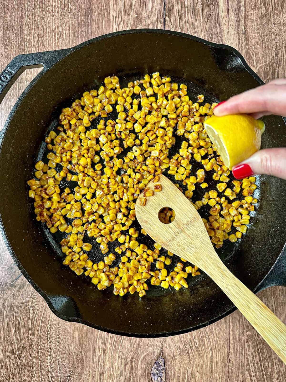 Hand squeezing a lemon in skillet with blackened corn and a wooden spoon.
