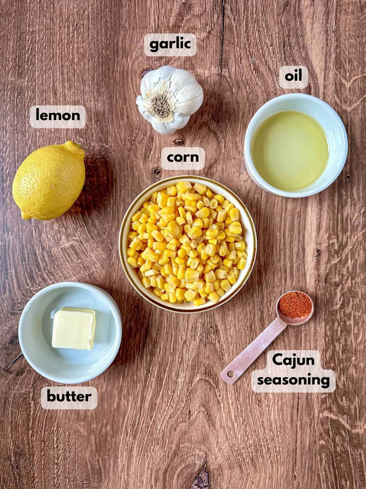 Ingredients need to make blackened corn on wooden table: corn kernels, garlic, butter, oil, lemon, and Cajun seasonings.