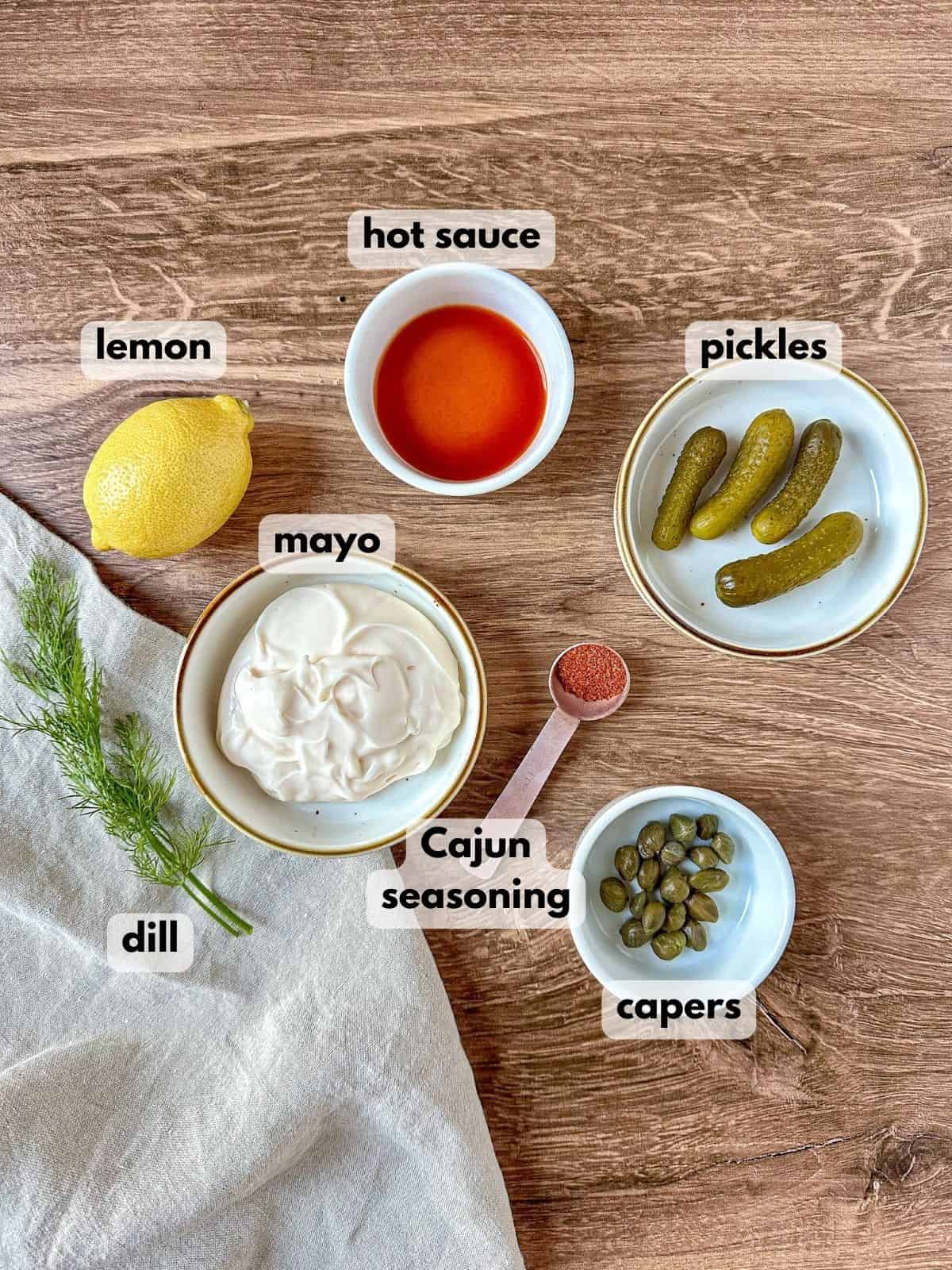 Cajun tartar sauce ingredients on a wooden table: mayo, pickles, capers, hot sauce, dill, seasonings, and a lemon.