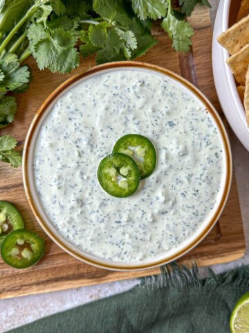 Creamy jalapeno sauce in a bowl surrounded by sliced jalapeno, cilantro, and tortilla chips.