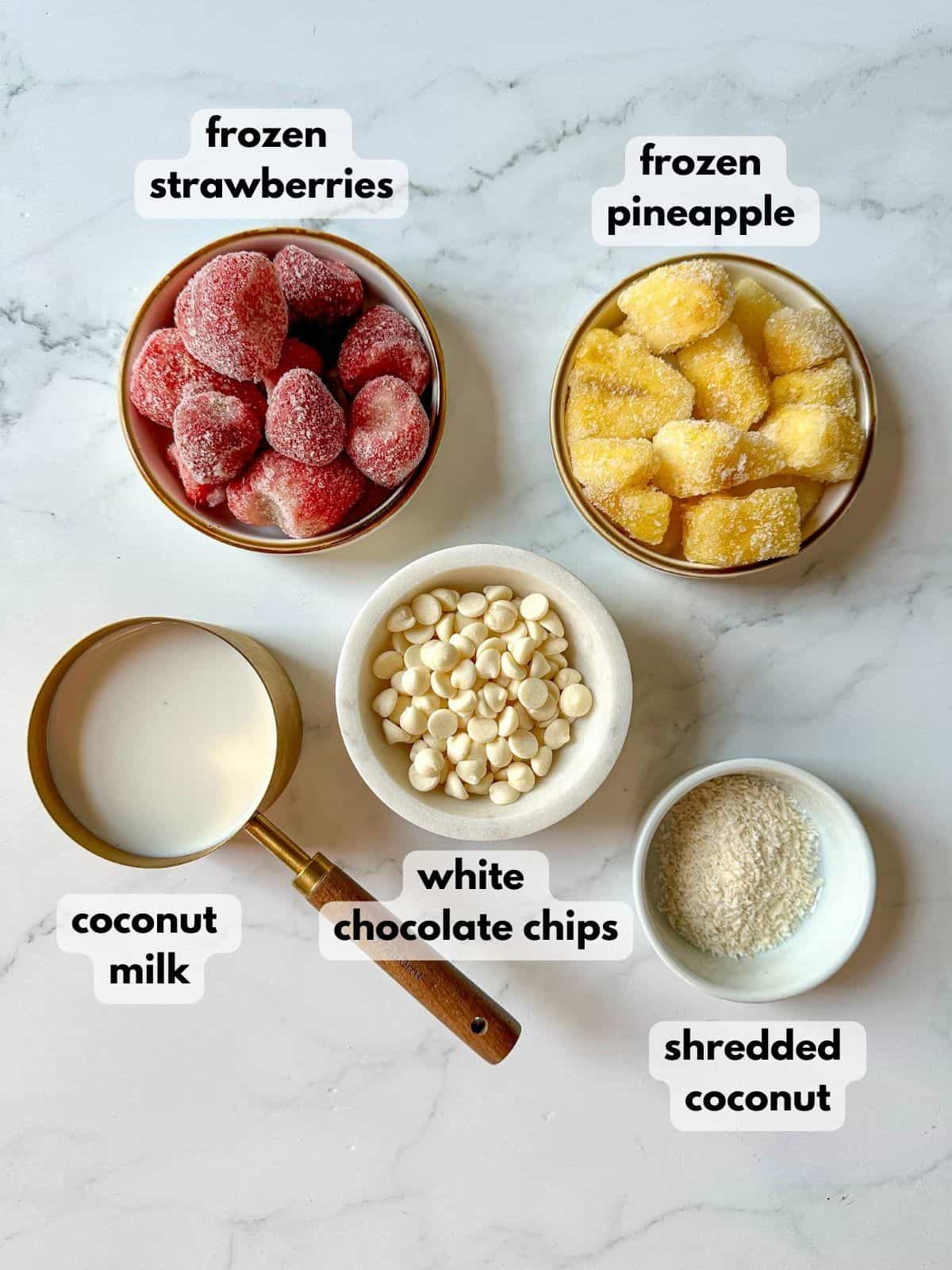 Frozen strawberries and pineapples in small bowls with coconut milk in a measuring cup and bowls of white chocolate and shredded coconut.