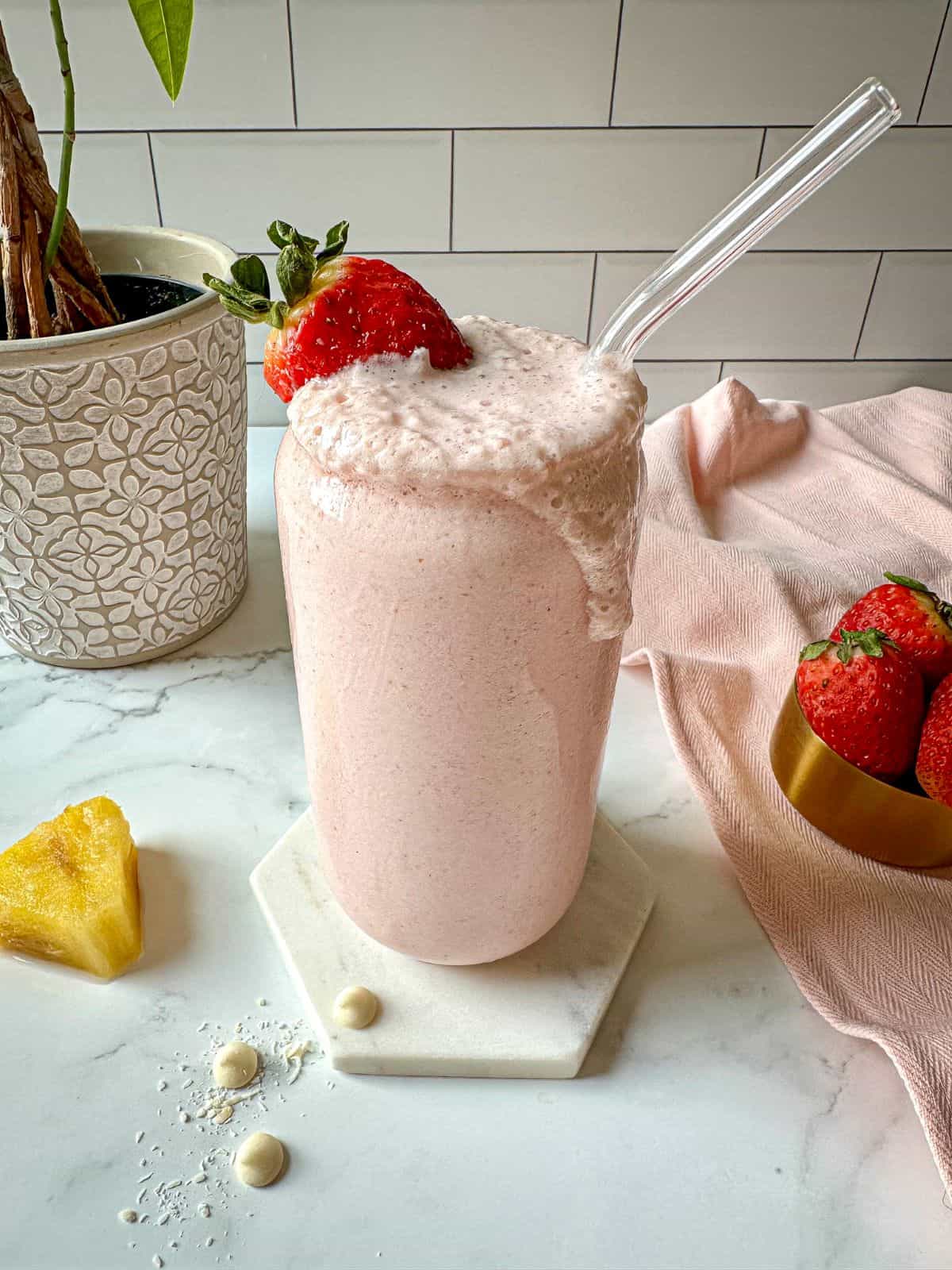 A pink Bahama mama tropical smoothie in a glass with a strawberry garnish on top. Pineapples, strawberries, white chocolate chips, and shredded coconut around the glass.