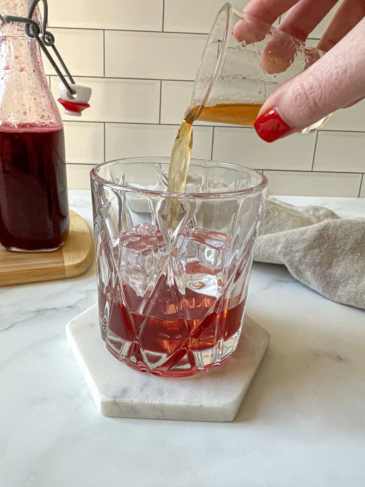 A hand pouring a jigger of bourbon into a cocktail glass filled with blackberry syrup and ice.