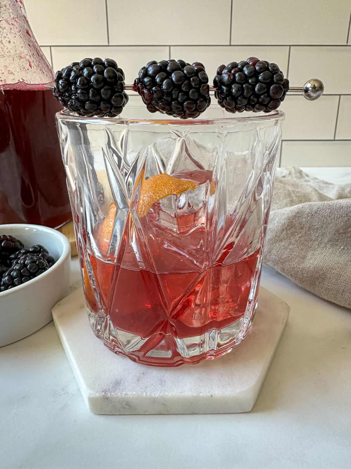 A purple blackberry old fashioned cocktail with a garnish of three blackberries and an orange peel.