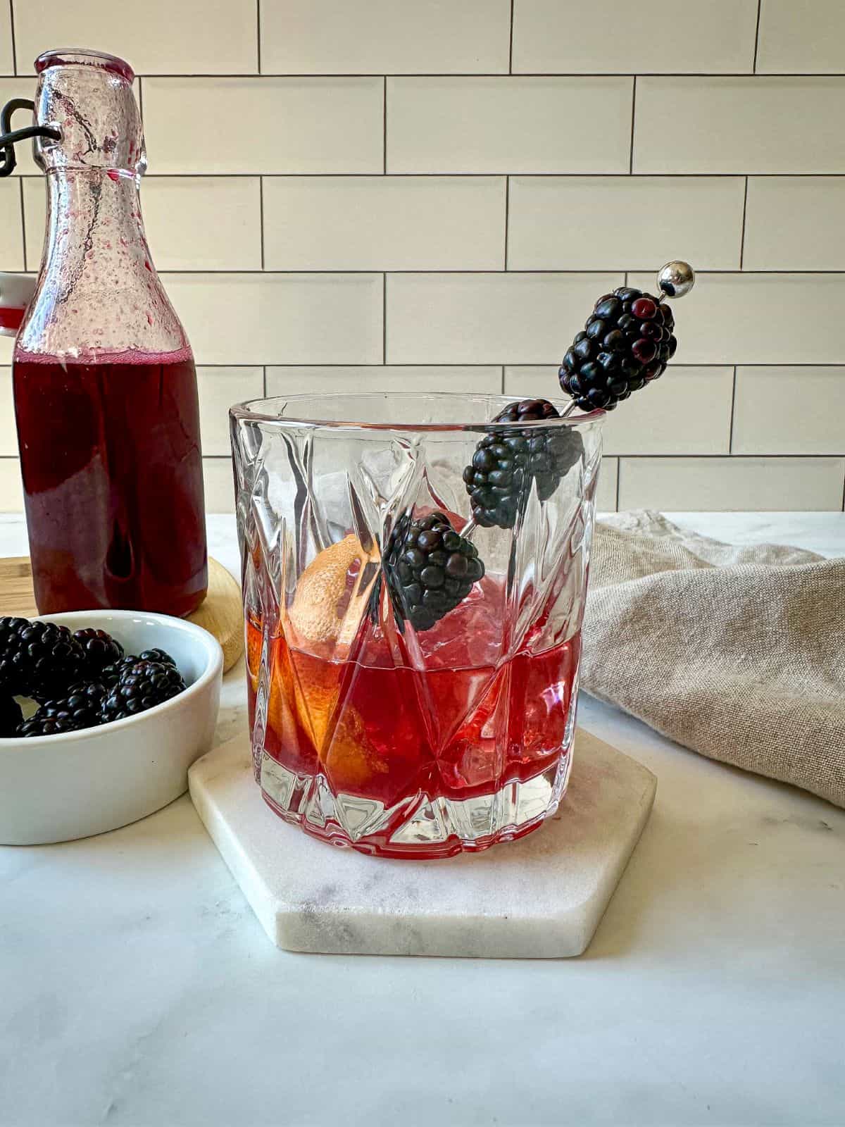 Blackberry old fashioned cocktail on a marble coaster with fresh blackberries and purple syrup in a glass jar.