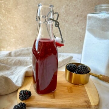 Glass jar filled with a purple blackberry simple syrup surrounded by fresh blackberries.