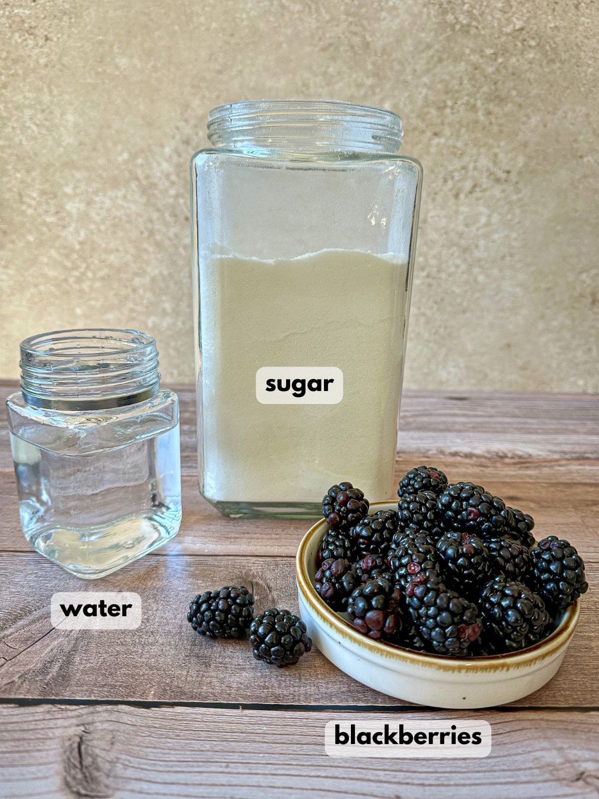 Fresh blackberries in a bowl, a glass container of sugar, and a glass of water.