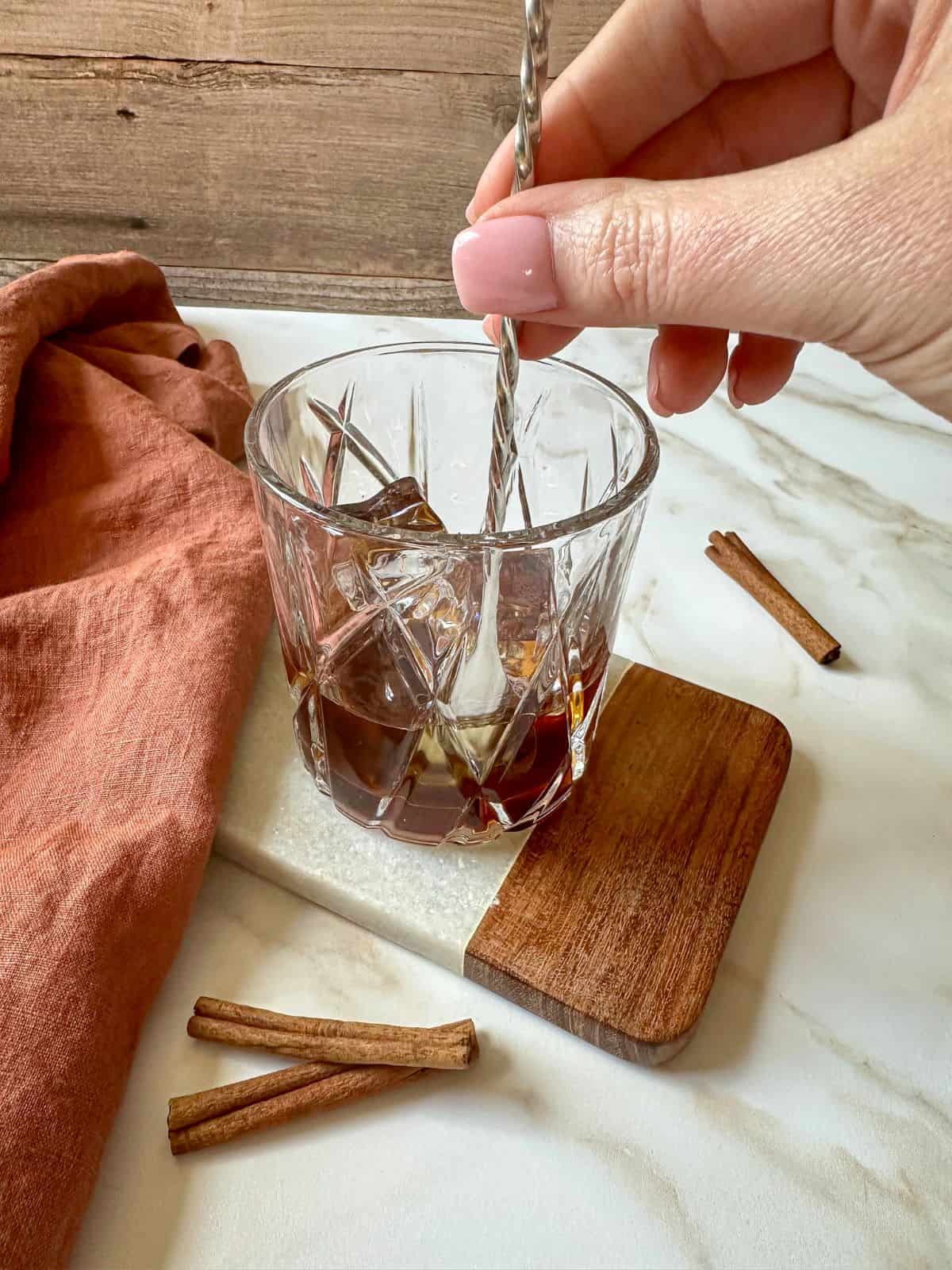 A hand using a bar spoon to stir the cinnamon old fashioned cocktail.