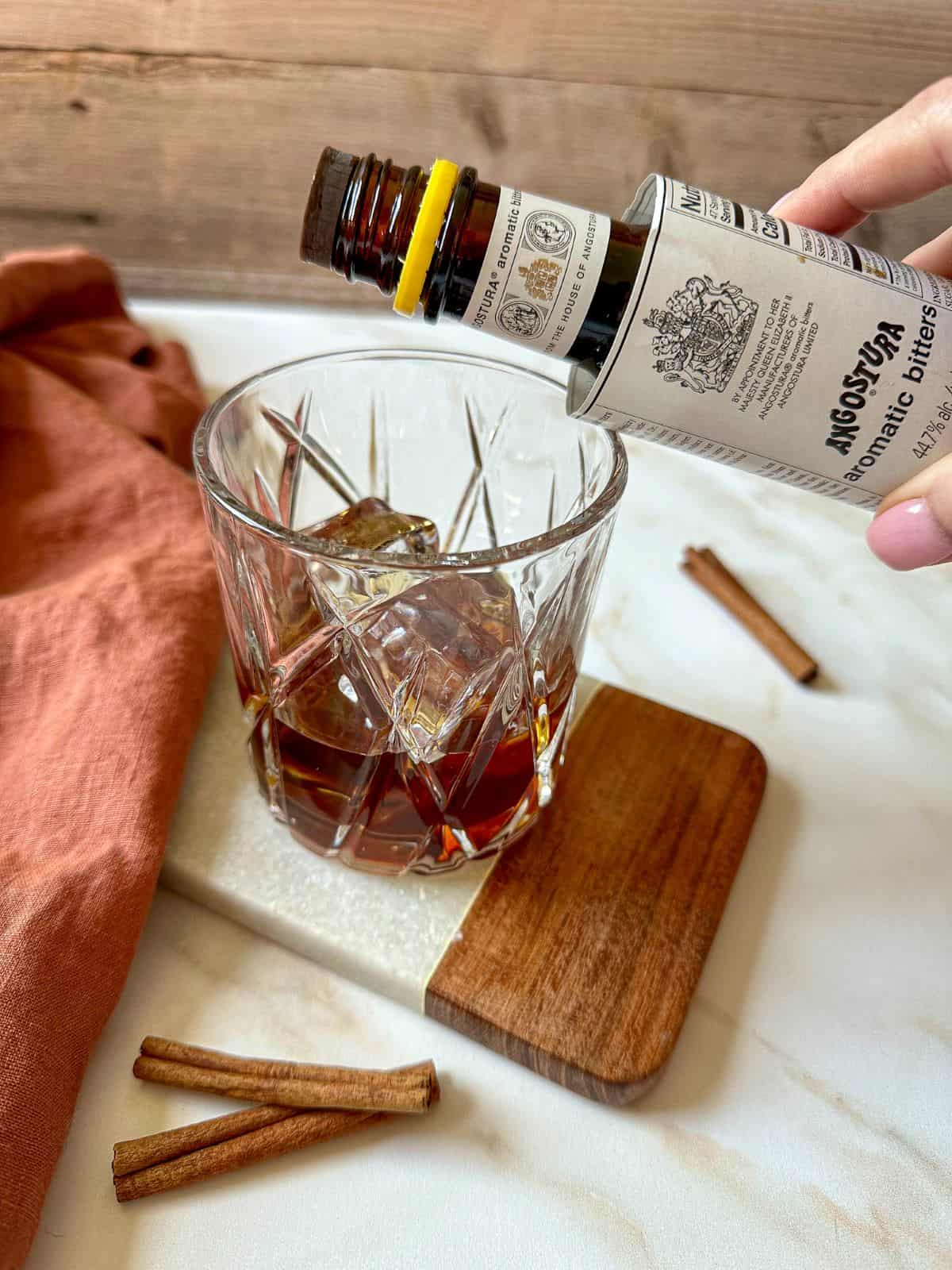 A hand holding an aromatic bitters bottle over a rocks glass filled with bourbon and cinnamon simple syrup.