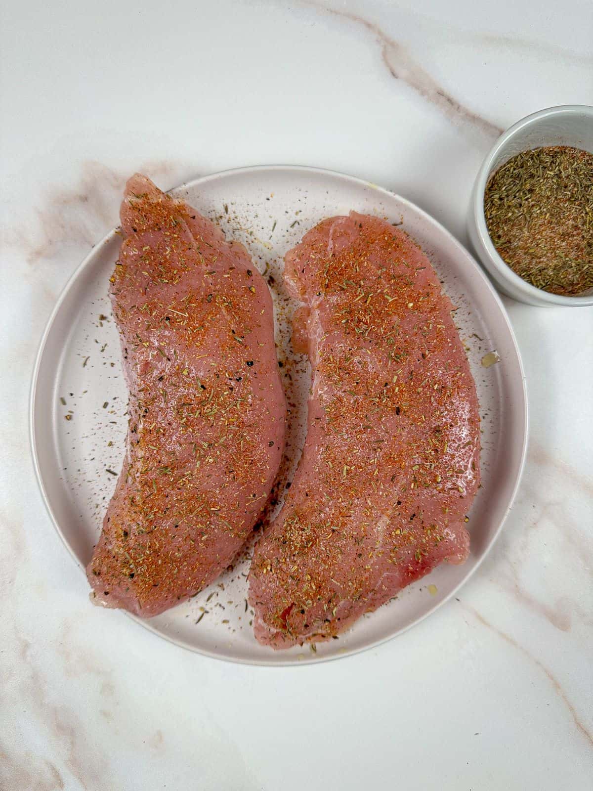 A plate of seasoned turkey breast fillets next to a bowl of seasonings.