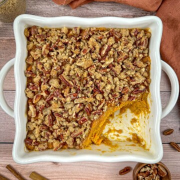 Old fashioned sweet potato casserole with pecans in a baking dish with a jar of brown sugar and cinnamon sticks on a wooden table.