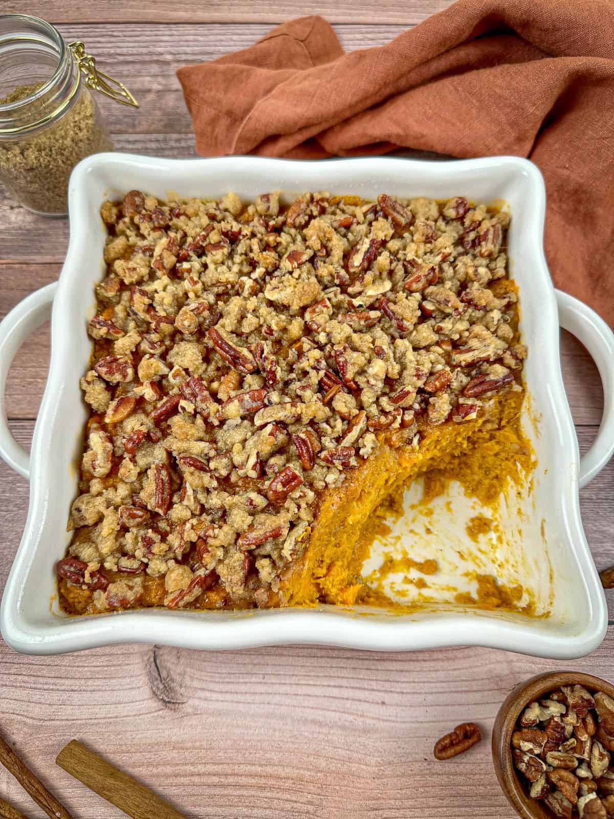 Bourbon sweet potato casserole with pecan topping in a baking dish with a jar of brown sugar and cinnamon sticks on a wooden table.