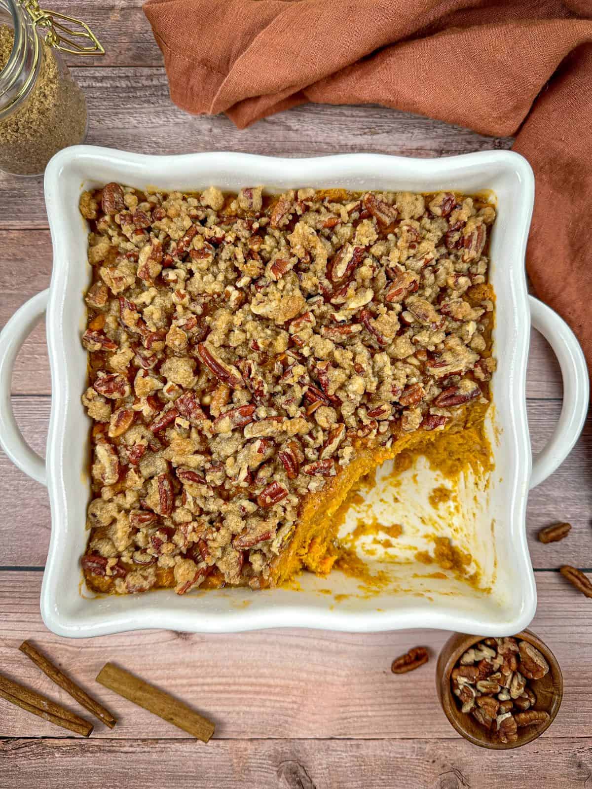 Old fashioned sweet potato casserole with pecans in a baking dish with a jar of brown sugar and cinnamon sticks on a wooden table.