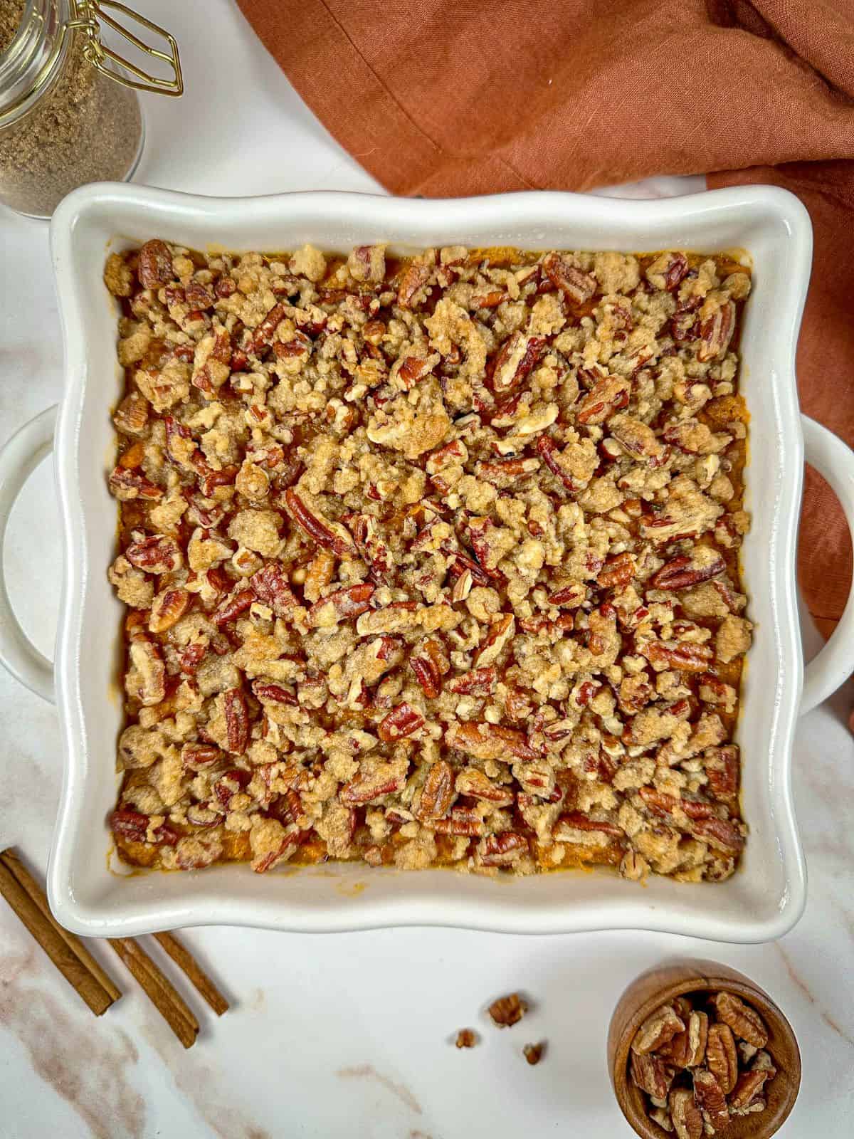 Old fashioned sweet potato casserole with pecan topping in a baking dish with a jar of brown sugar and cinnamon sticks on a marble countertop.