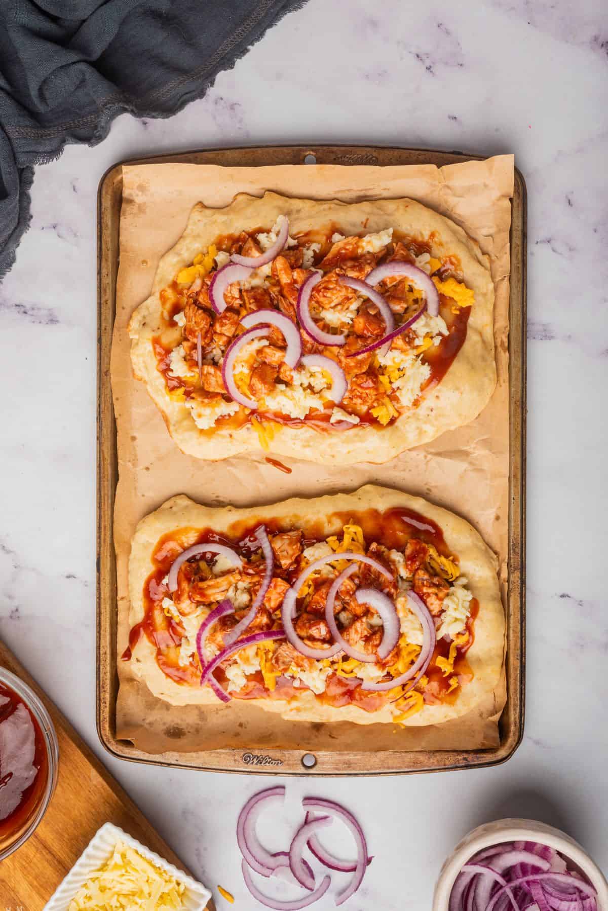 Two flatbread crusts on a baking tray with bbq sauce, chicken, red onions, and cheese.