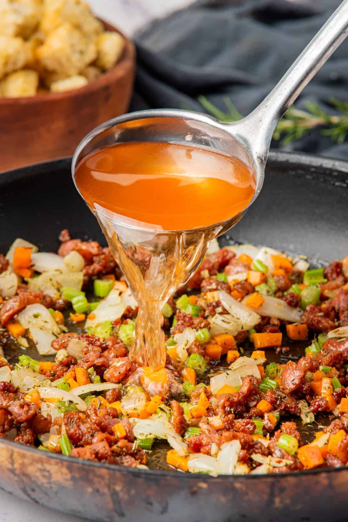 A ladle pouring chicken broth into the skillet with ground Italian sausage and sauteed vegetables.