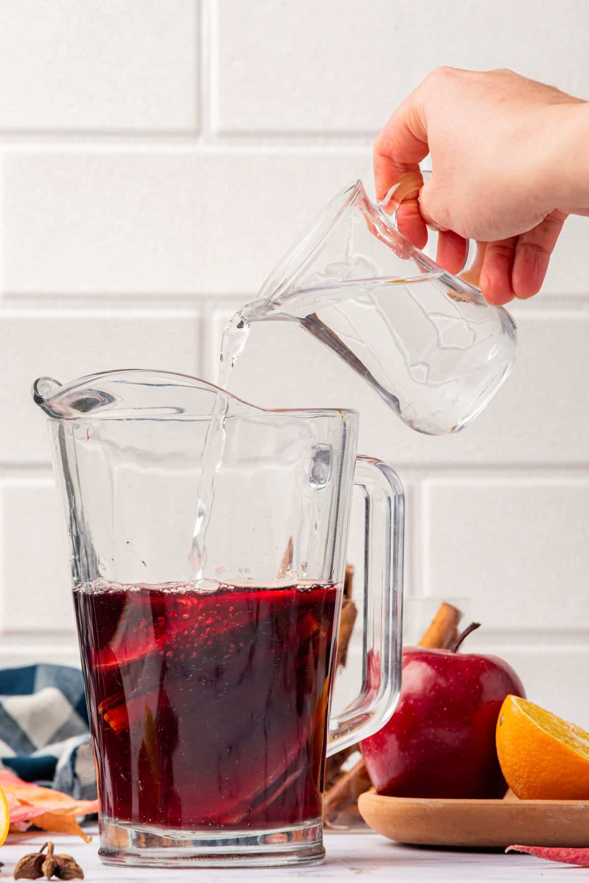 Hand pouring Cointreau in a pitcher of red wine sangria with cinnamon sticks and apple slices.