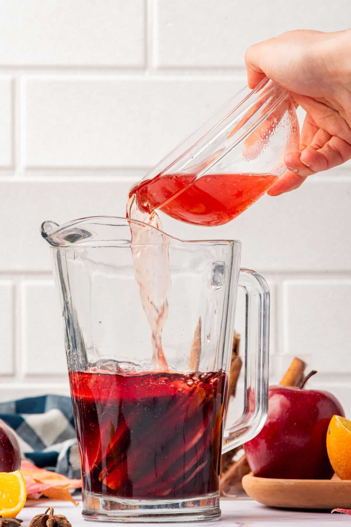 Hand pouring cranberry juice into a pitcher of red wine and cinnamon sticks and apple slices.