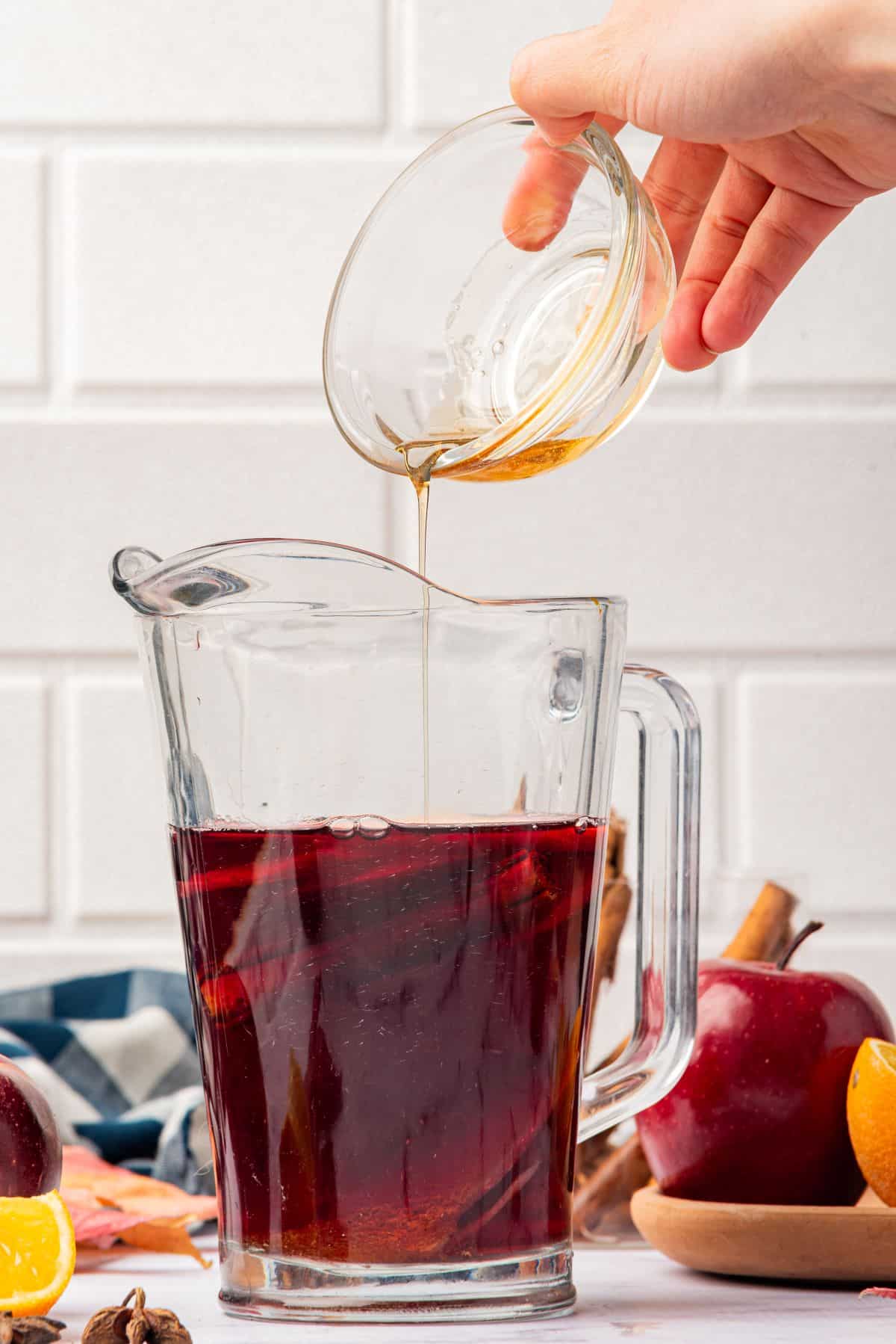 Hand pouring maple syrup in a pitcher of Thanksgiving sangria with cranberry juice and fall ingredients.