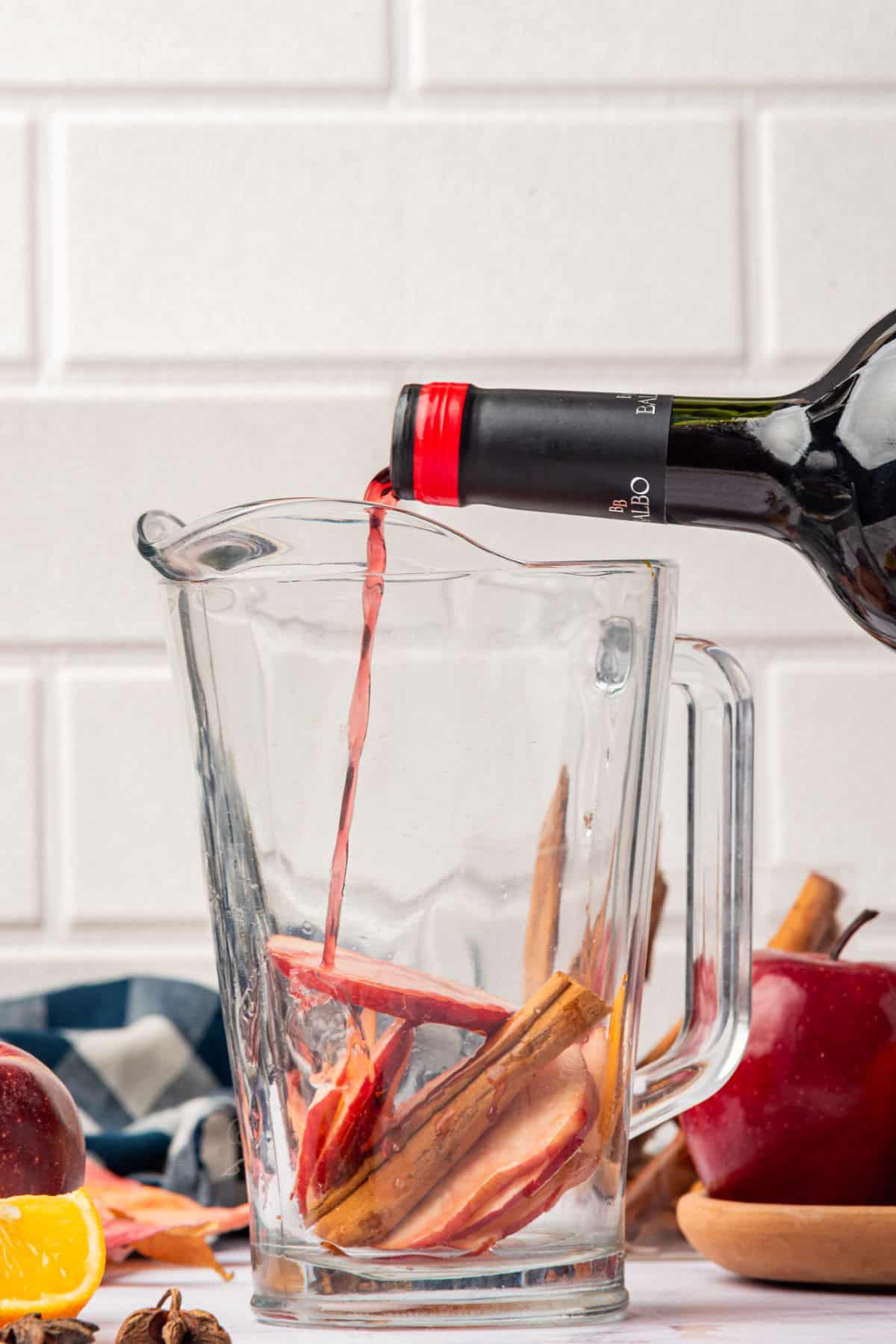 Pouring red wine into a large pitcher with sliced apples and cinnamon sticks.