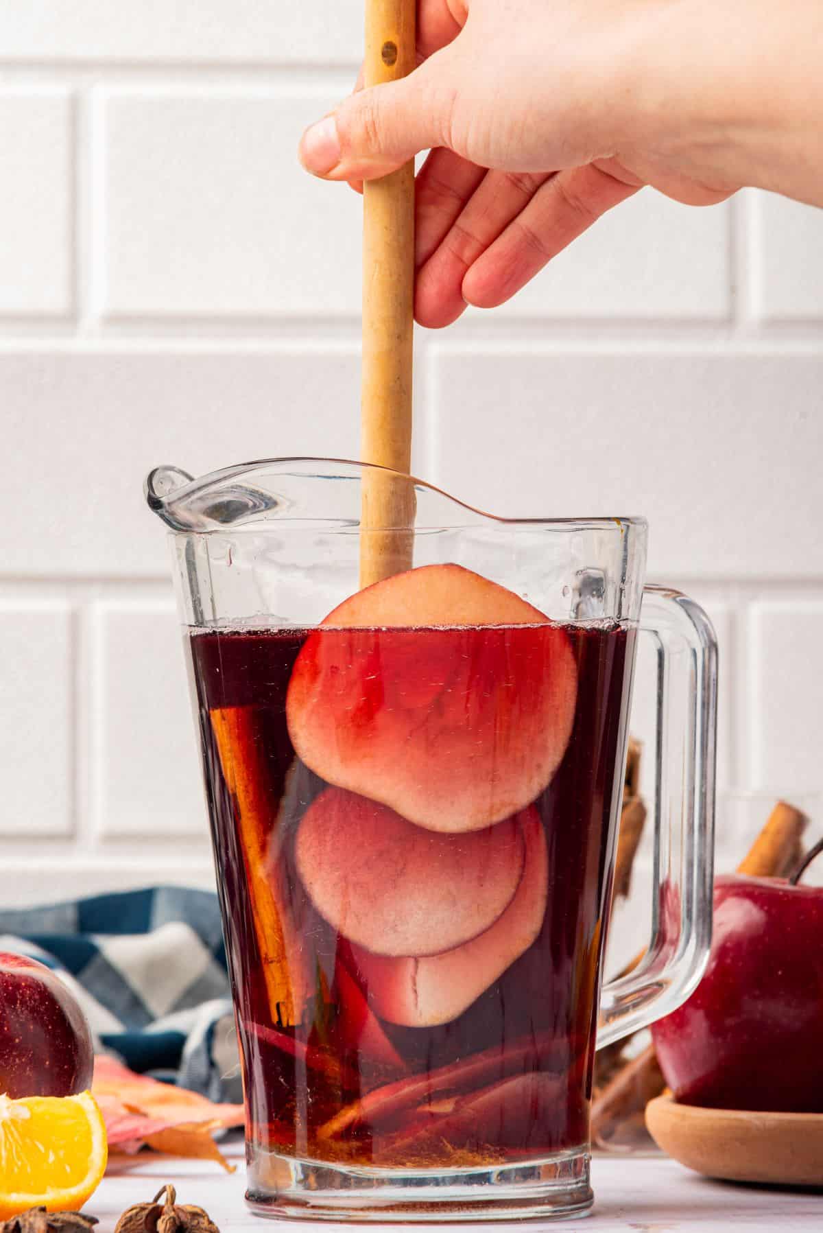 Stirring a pitcher of Thanksgiving red sangria with cranberry juice, cinnamon sticks, and fruit with a wooden spoon.