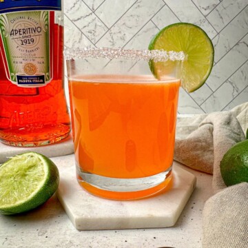 Aperol margarita, an orange drink, in a rocks glass with a salt rim and lime garnish.