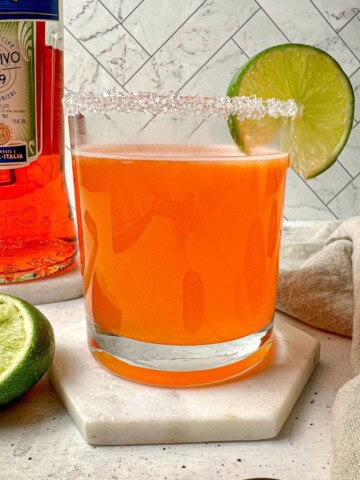 Aperol margarita, an orange drink, in a rocks glass with a salt rim and lime garnish.