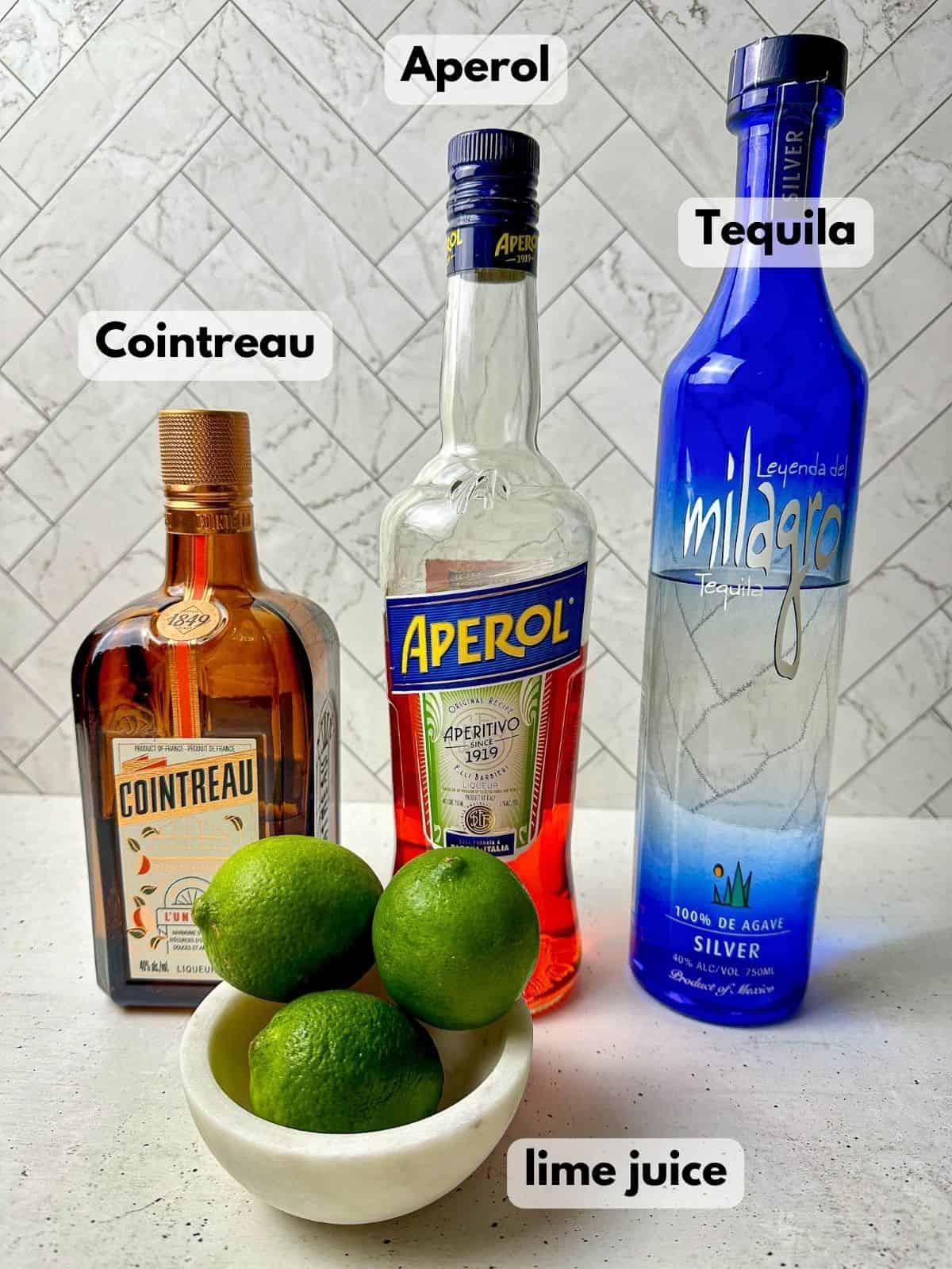 Aperol margarita ingredients include bottles of Aperol, tequila, Cointreau, and a bowl of limes.