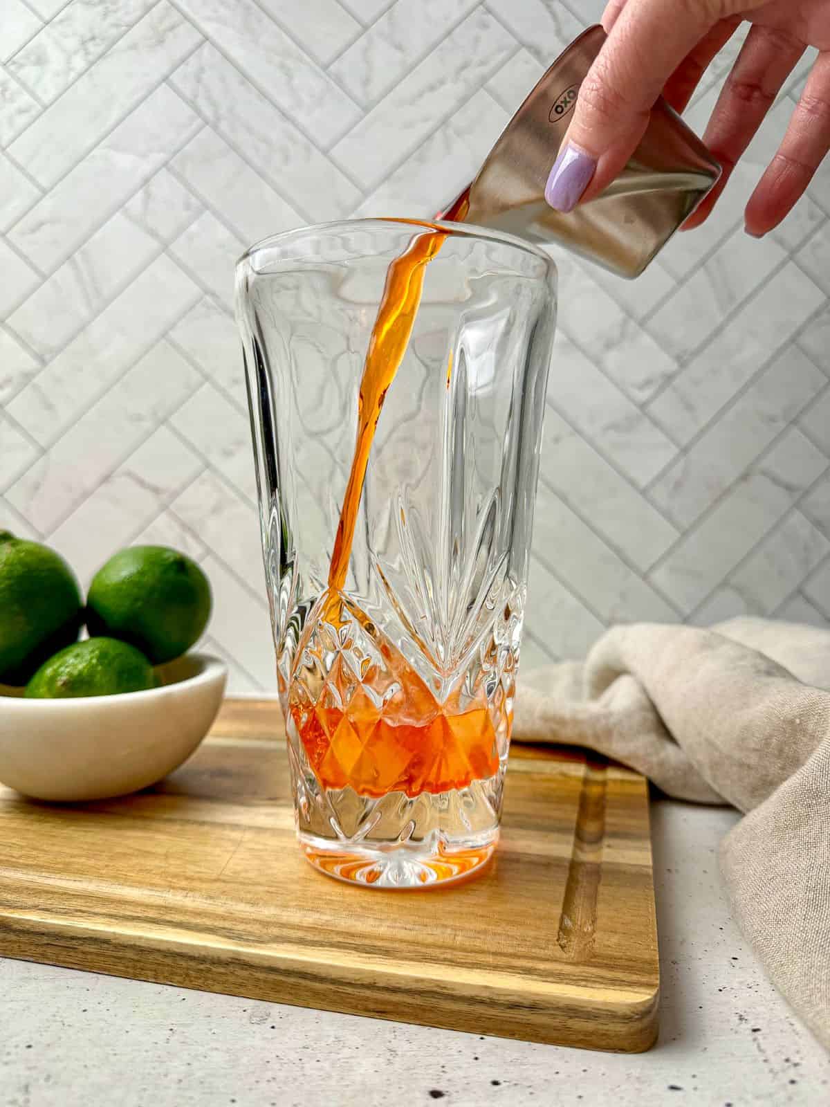 A hand pouring Aperol into a cocktail shaker with ice.