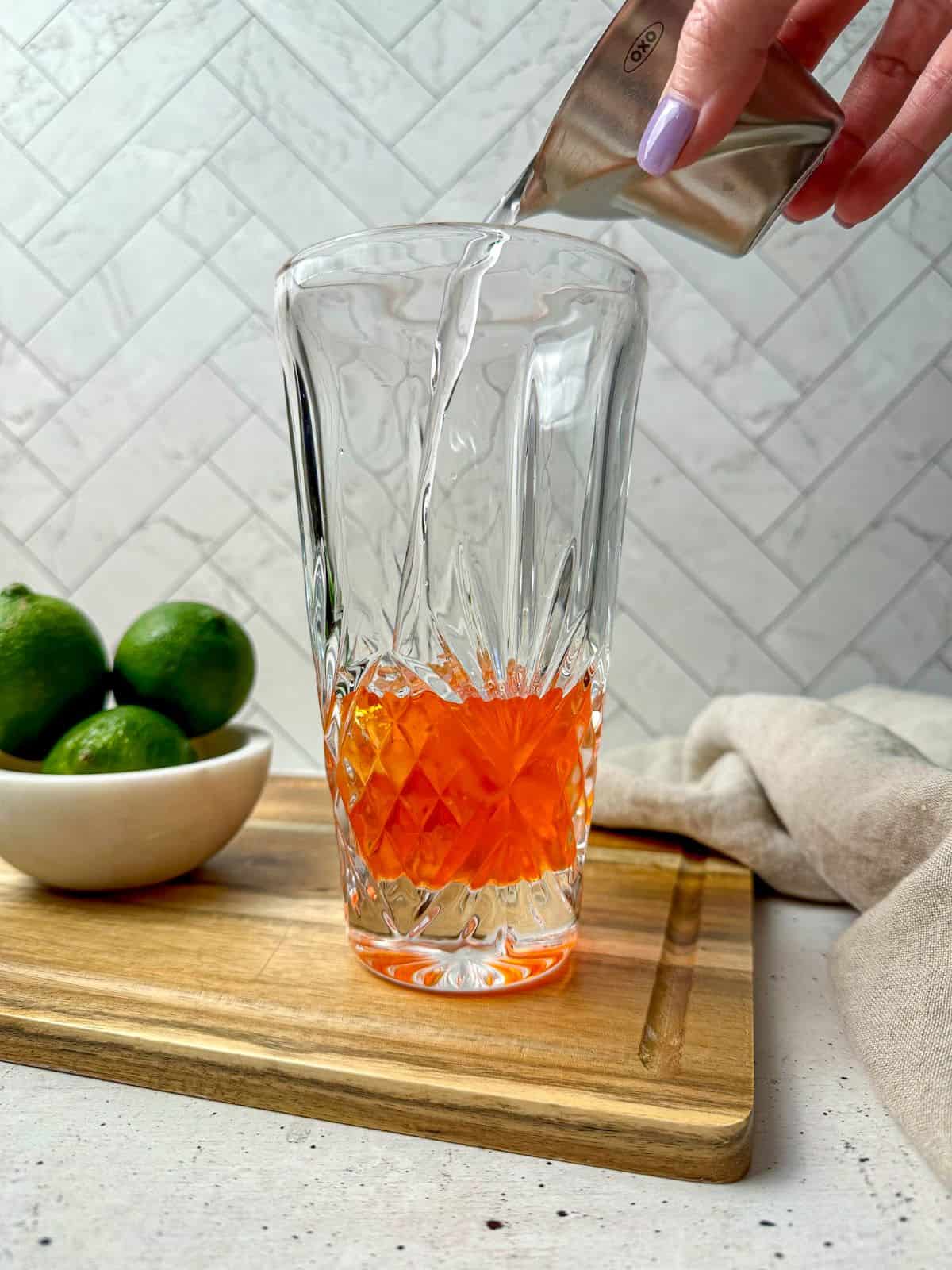 A hand pouring tequila into a cocktail shaker with Aperol.