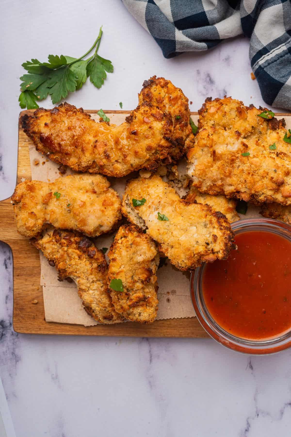 Crispy Cheez-It chicken tenders on a wooden cutting board with a bowl of ketchup for dipping.