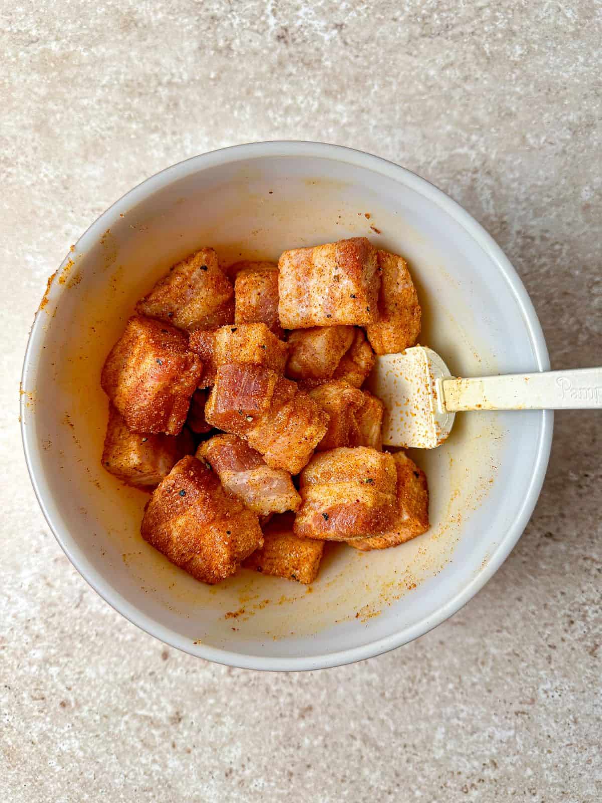 A spoon in a mixing bowl with seasoned pork belly cubes.