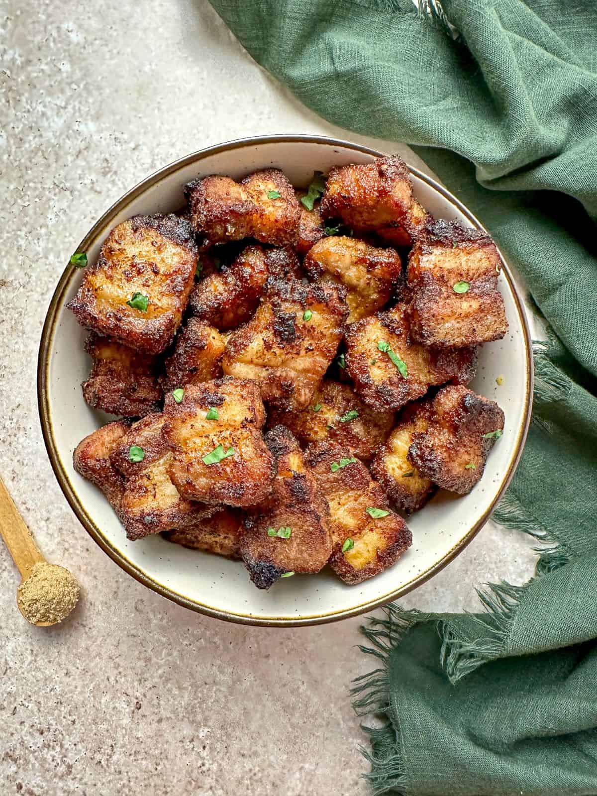 Air fried pork belly bites in a serving bowl with a teaspoon of brown sugar  and green towel on the countertop.