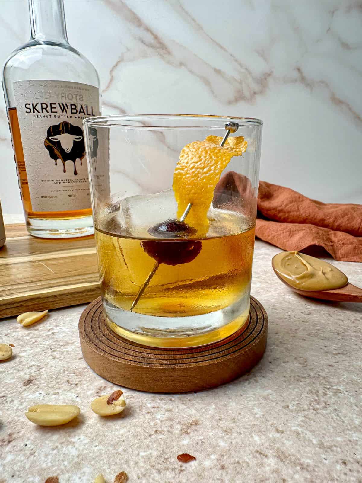 Peanut butter old fashioned with orange peel and cherry garnish with a bottle of Skrewball whiskey on a cutting board.