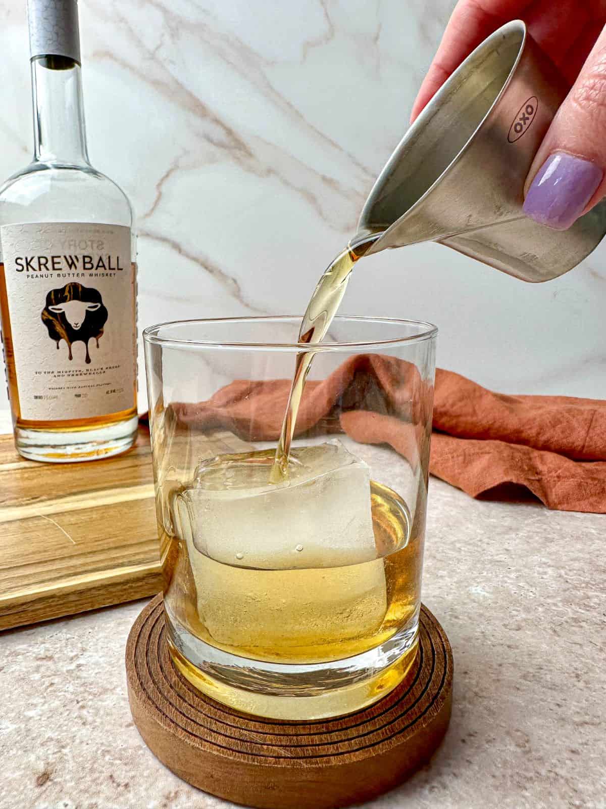 Pouring Skrewball peanut butter whiskey in a lowball glass with a large ice cube.