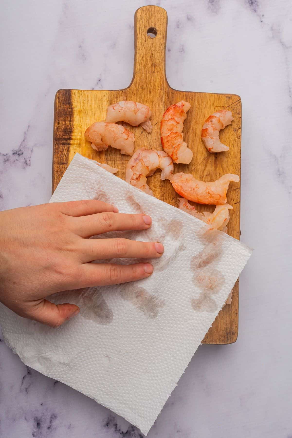 A hand patting shrimp dry with a paper towel on a wooden cutting board.