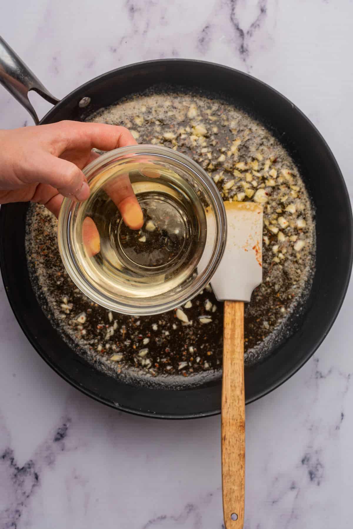 A hand holding a bowl of white wine over a skillet with garlic sauteing in butter and olive oil.